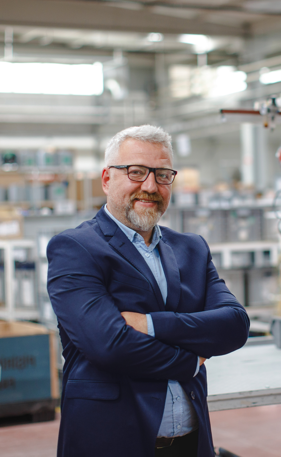 Smiling middle-aged man with gray hair and beard, wearing glasses and a navy blazer, standing with arms crossed in an industrial or warehouse setting.