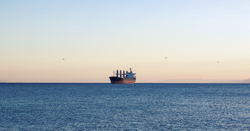 Cargo ship sailing on calm blue ocean waters at sunset with a clear sky and distant mountains.