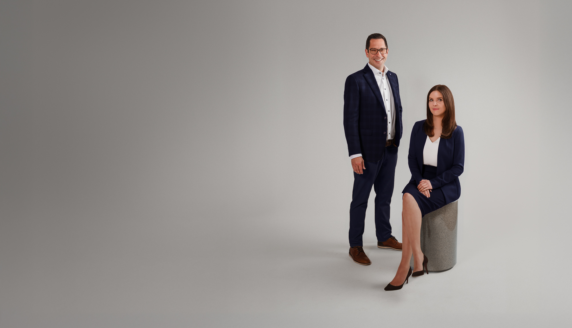 A man in a dark blue suit and glasses stands next to a woman in a matching dark blue suit sitting on a gray cylindrical stool against a light gray background.
