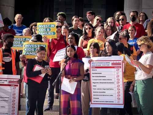Photo of Aparna speaking into a microphone while wearing a multicolor jumpsuit, with a crowd of people standing behind her