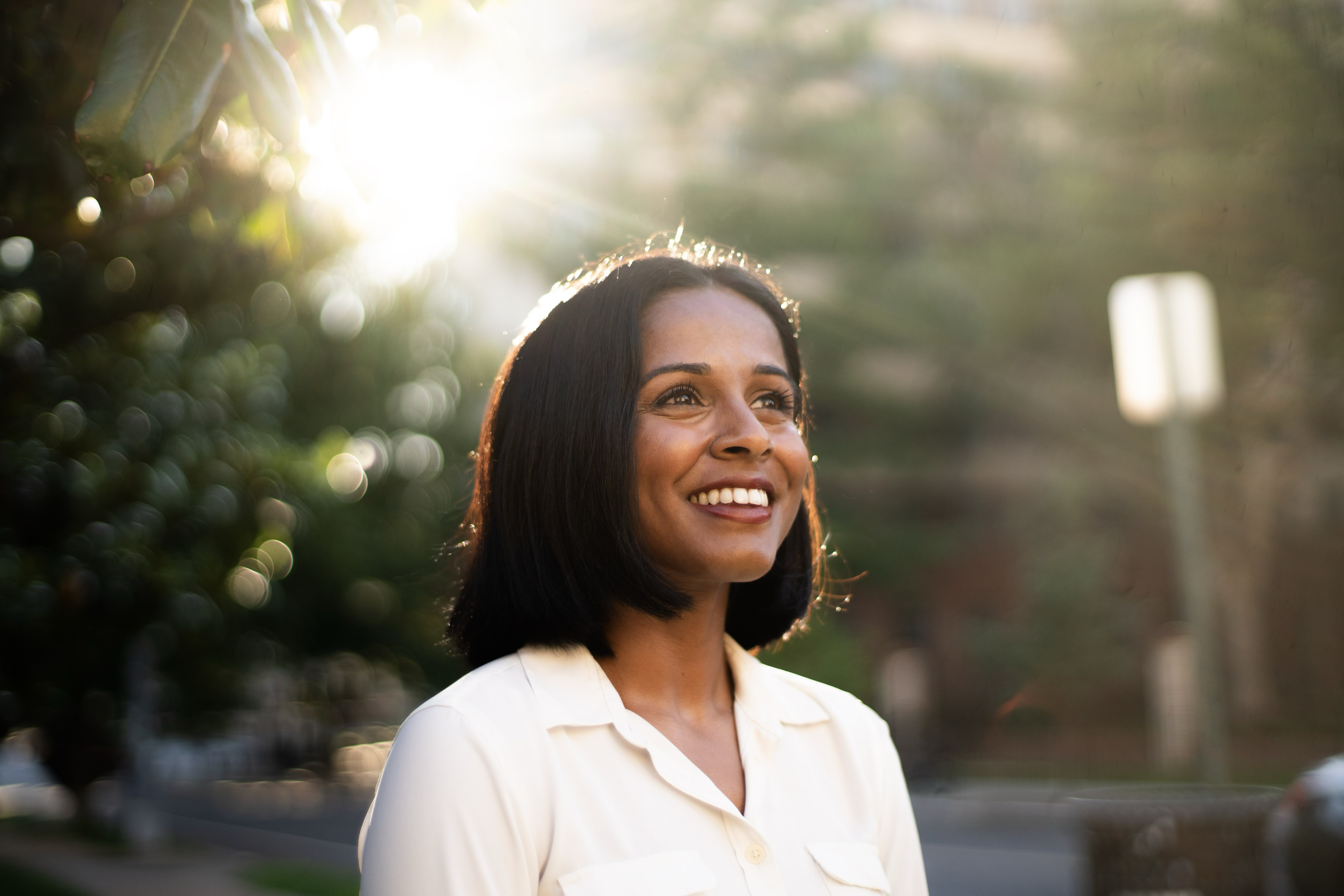 Photo of Aparna smiling and looking upwards. She is wearing a white button down and there are trees and the sun shining behind her