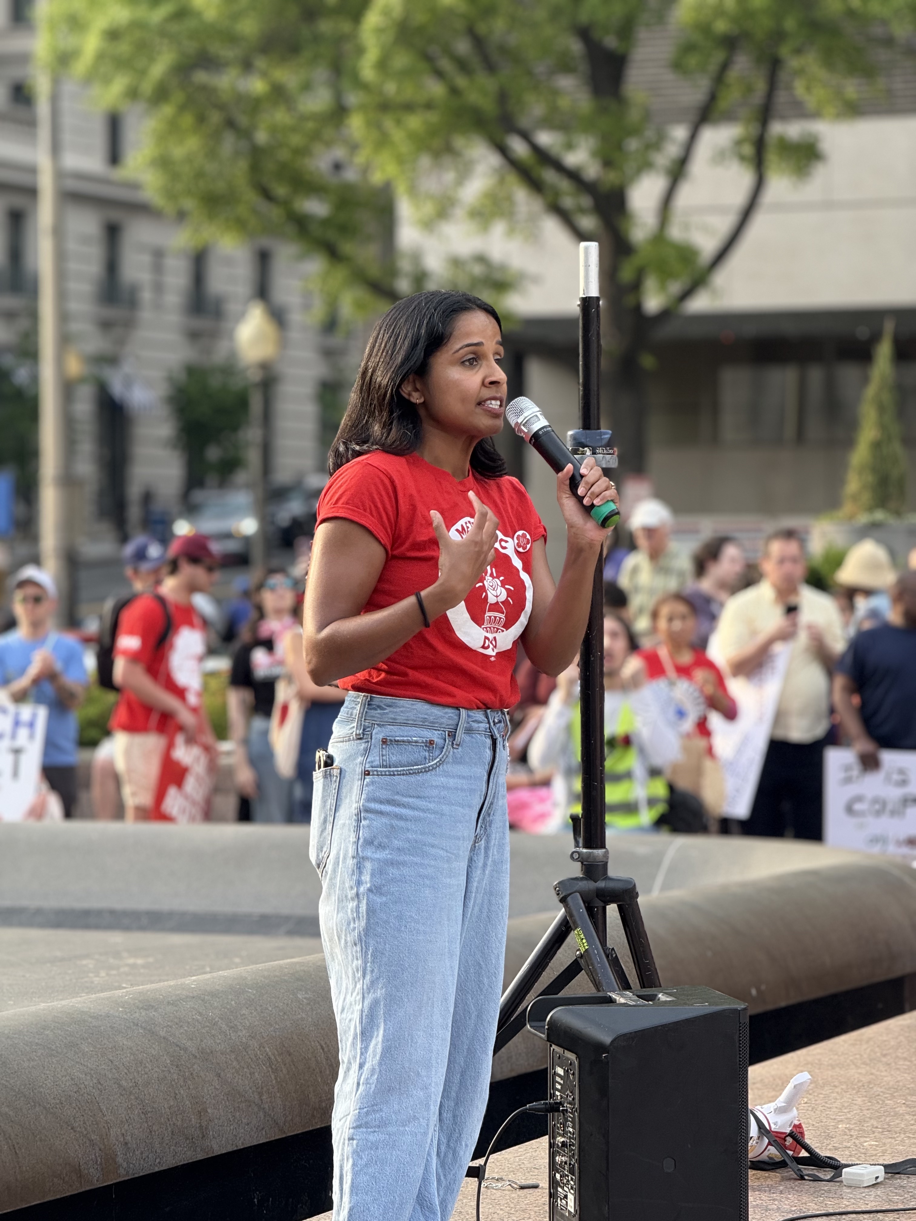 Photo of Aparna wearing a Metro DC DSA tshirt while speaking into a microphone with a crowd behind her