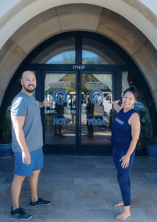 Smiling man and woman in athletic wear gesturing toward glass doors of The Foundry Hot Yoga studio.