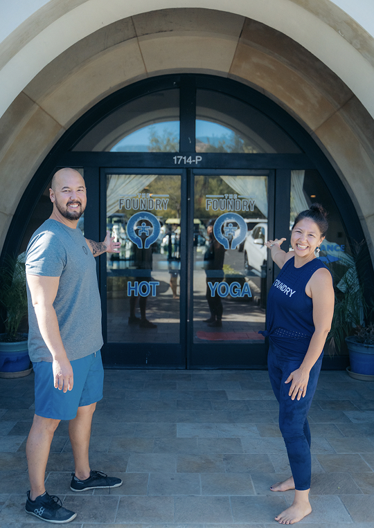 Smiling man and woman in athletic wear gesturing toward glass doors of The Foundry Hot Yoga studio.