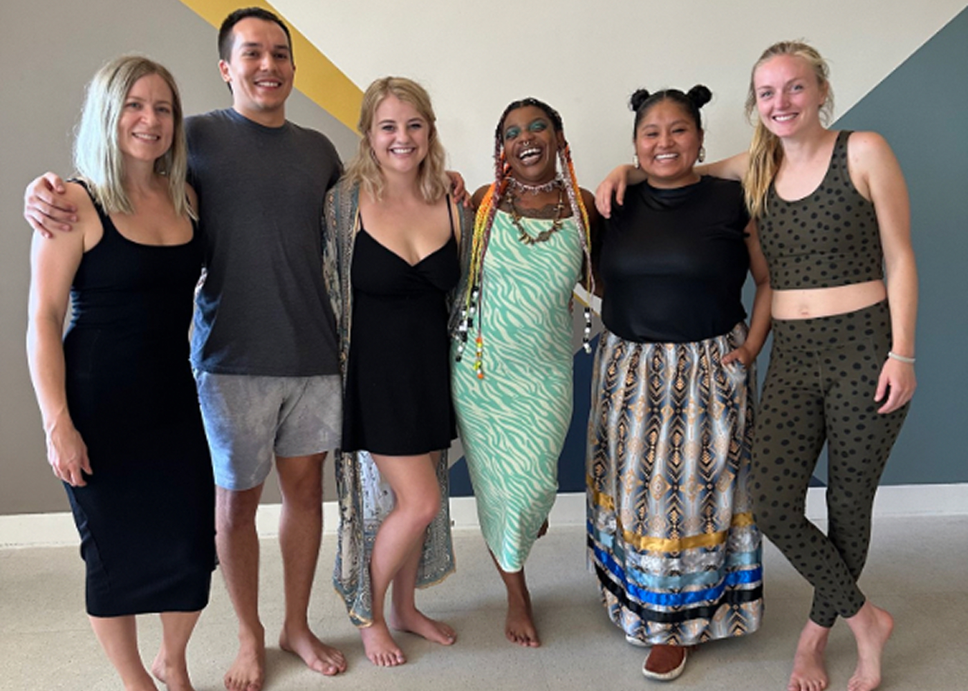 Group of six diverse young adults standing barefoot indoors smiling with arms around each other.