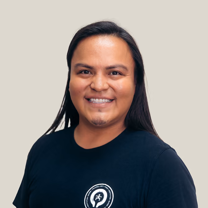 Smiling man with long dark hair wearing a black t-shirt with a circular logo, set against a plain light background.