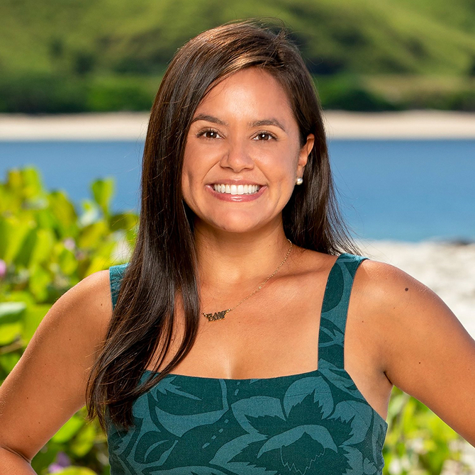 Smiling woman with long dark hair wearing a green patterned sleeveless top standing outdoors with greenery and water in the background.