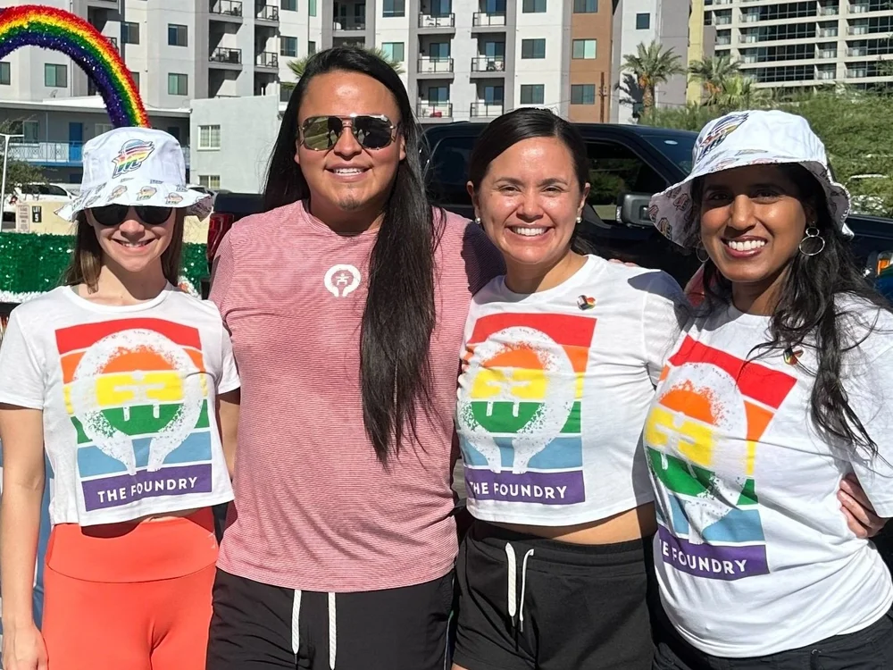 Group of four smiling people standing outdoors, three wearing white T-shirts with rainbow 'The Foundry' logo and two wearing white bucket hats.