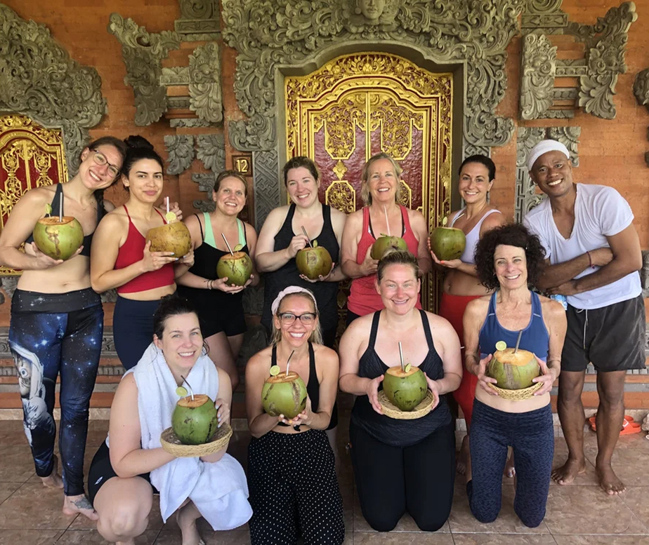 Group of eleven people smiling and holding coconuts with straws in front of ornate golden and stone carved doors.