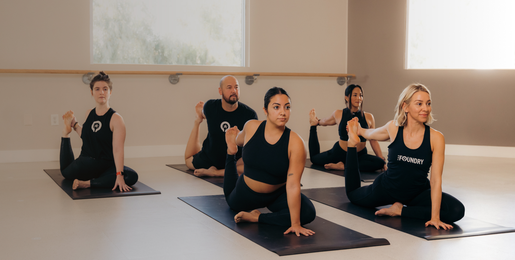 Five people practicing yoga indoors on mats, performing a seated pose with one leg bent and holding their foot with one hand.