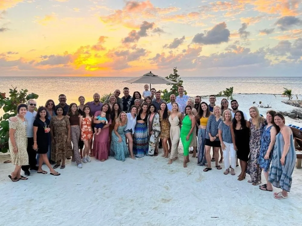 Large group of people posing on a beach at sunset with ocean and partly cloudy sky in the background.