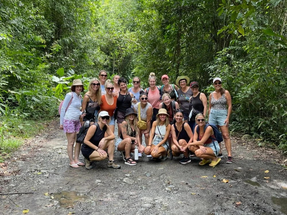 Group of 17 women posing on a forest trail surrounded by dense green foliage, dressed in casual summer clothes and hats.