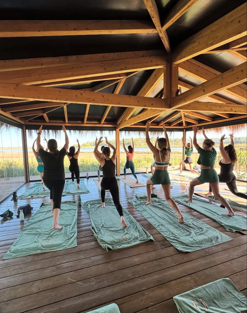 Group of people practicing yoga inside a wooden pavilion with large windows overlooking greenery and water.