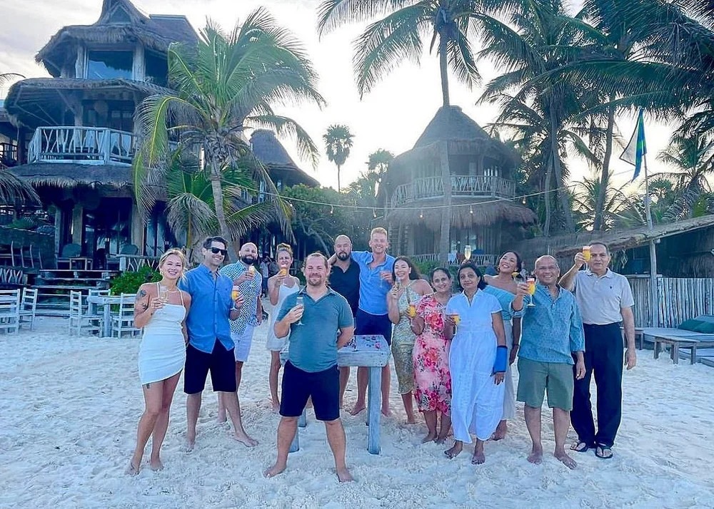 Group of barefoot people standing on sandy beach holding drinks, with tropical palm trees and thatched-roof huts in the background.
