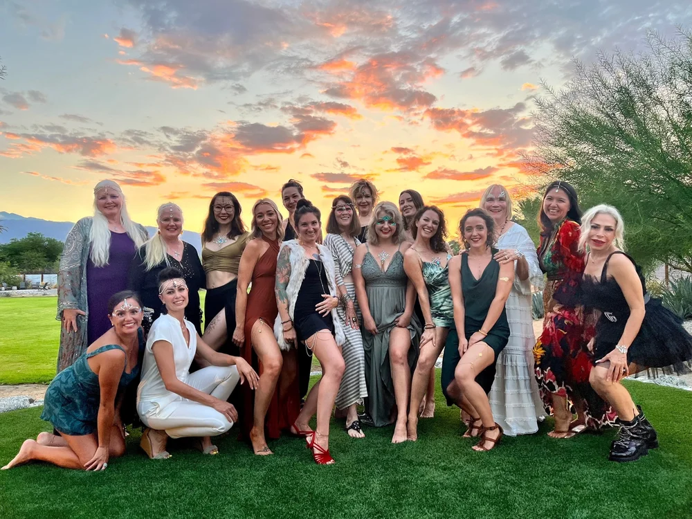 Group of fourteen women posing on green grass at sunset with colorful clouds, dressed in festive attire with decorative face gems.