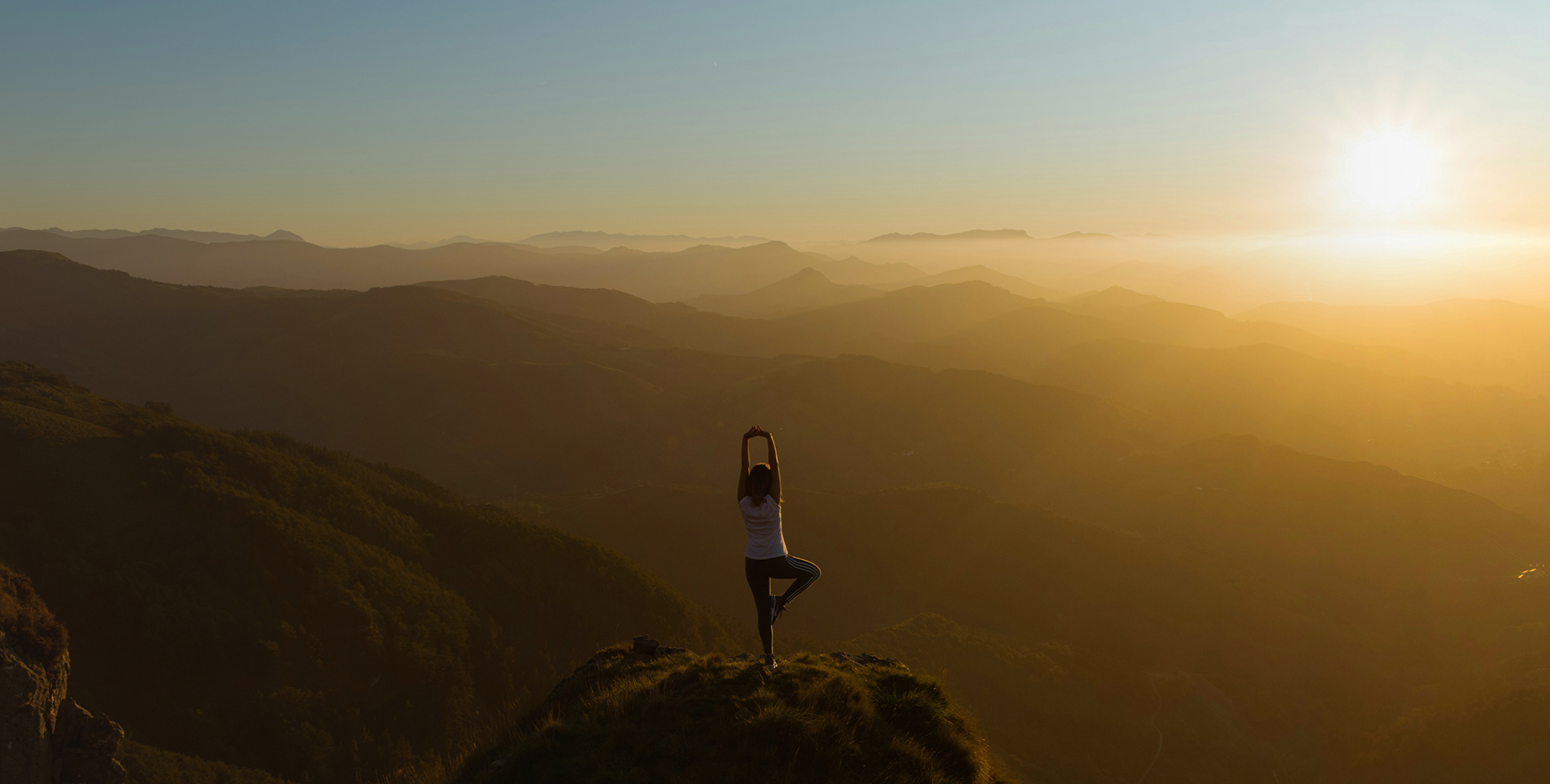 Person practicing yoga in tree pose on a mountain peak at sunrise with misty mountain layers in the background.