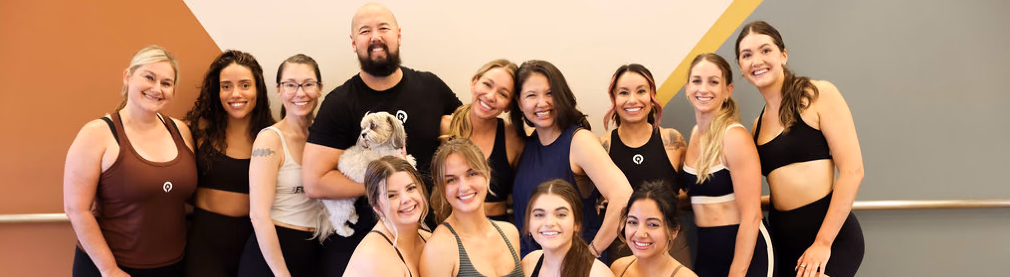 Group of smiling people in fitness attire posing together indoors with a small dog held by a man.