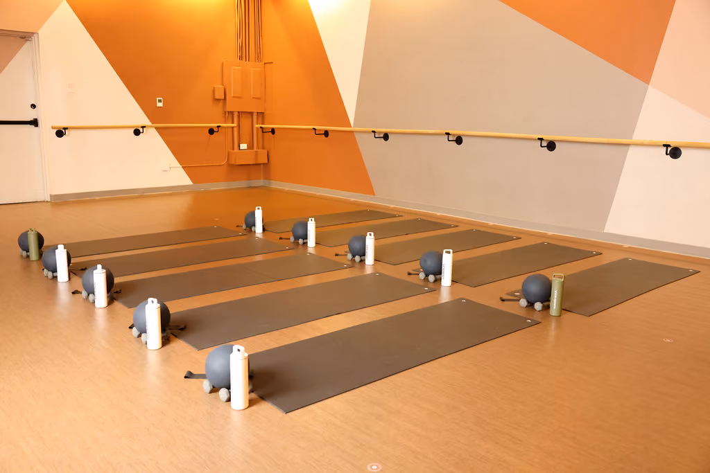 Yoga studio with six gray mats, exercise balls, small weights, and water bottles on wood floor.