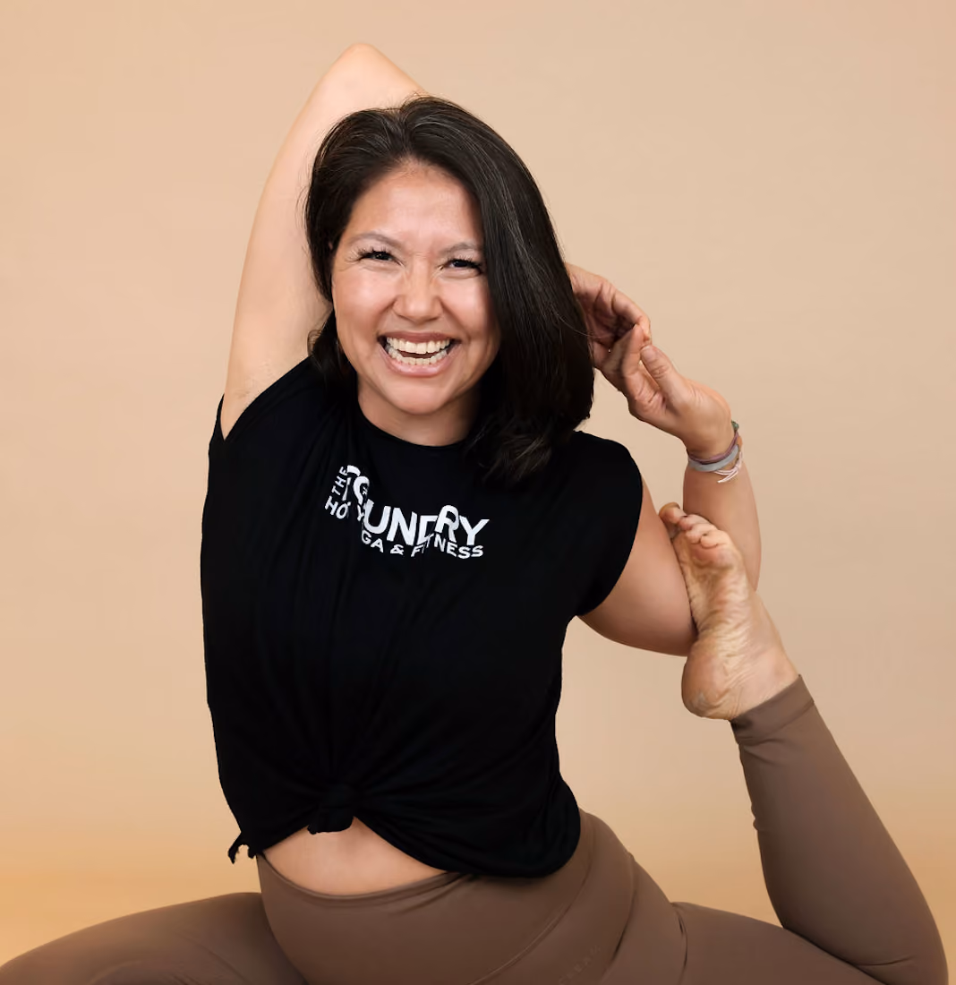 Smiling woman in black shirt and brown leggings doing a yoga stretch with a beige background.