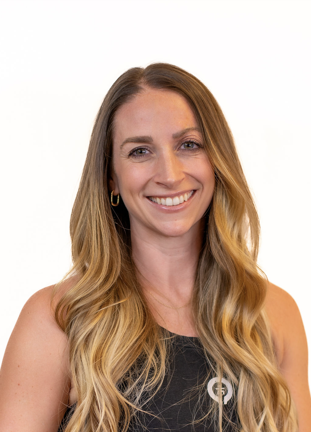 Smiling woman with long wavy blonde hair wearing a dark sleeveless top against a white background.