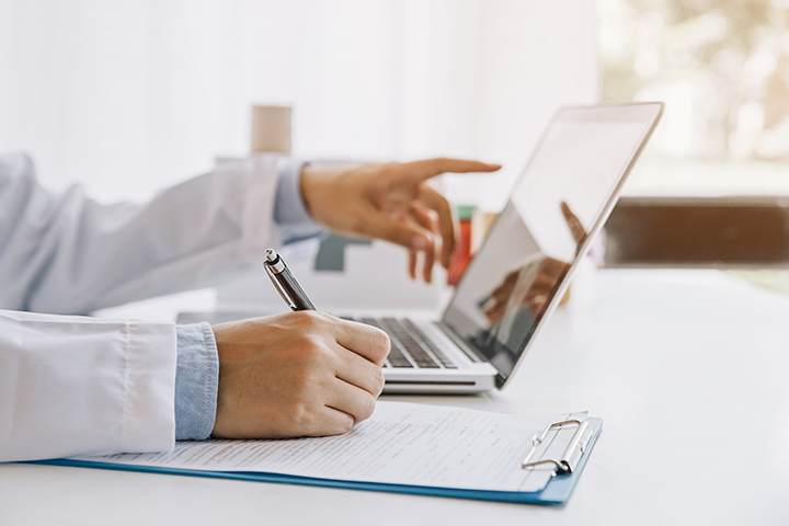 Un ordinateur portable vu de latérale, avec deux mains : l’une écrit au clavier, l’autre pointe l’écran.