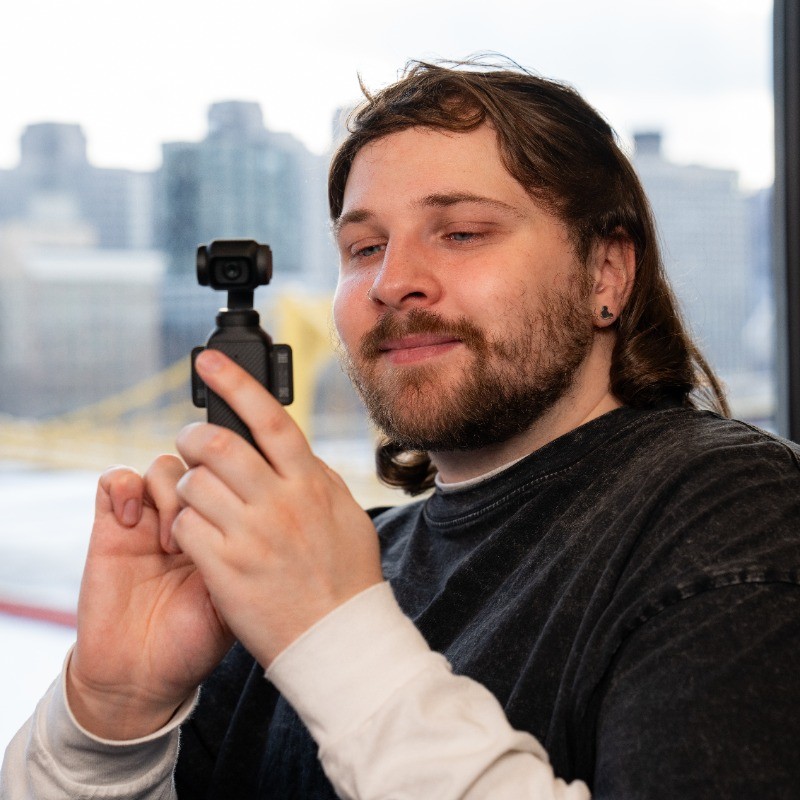 Logan LaMaster holding a camera in his hand as he takes video footage. The pittsburgh bridge is behind him.