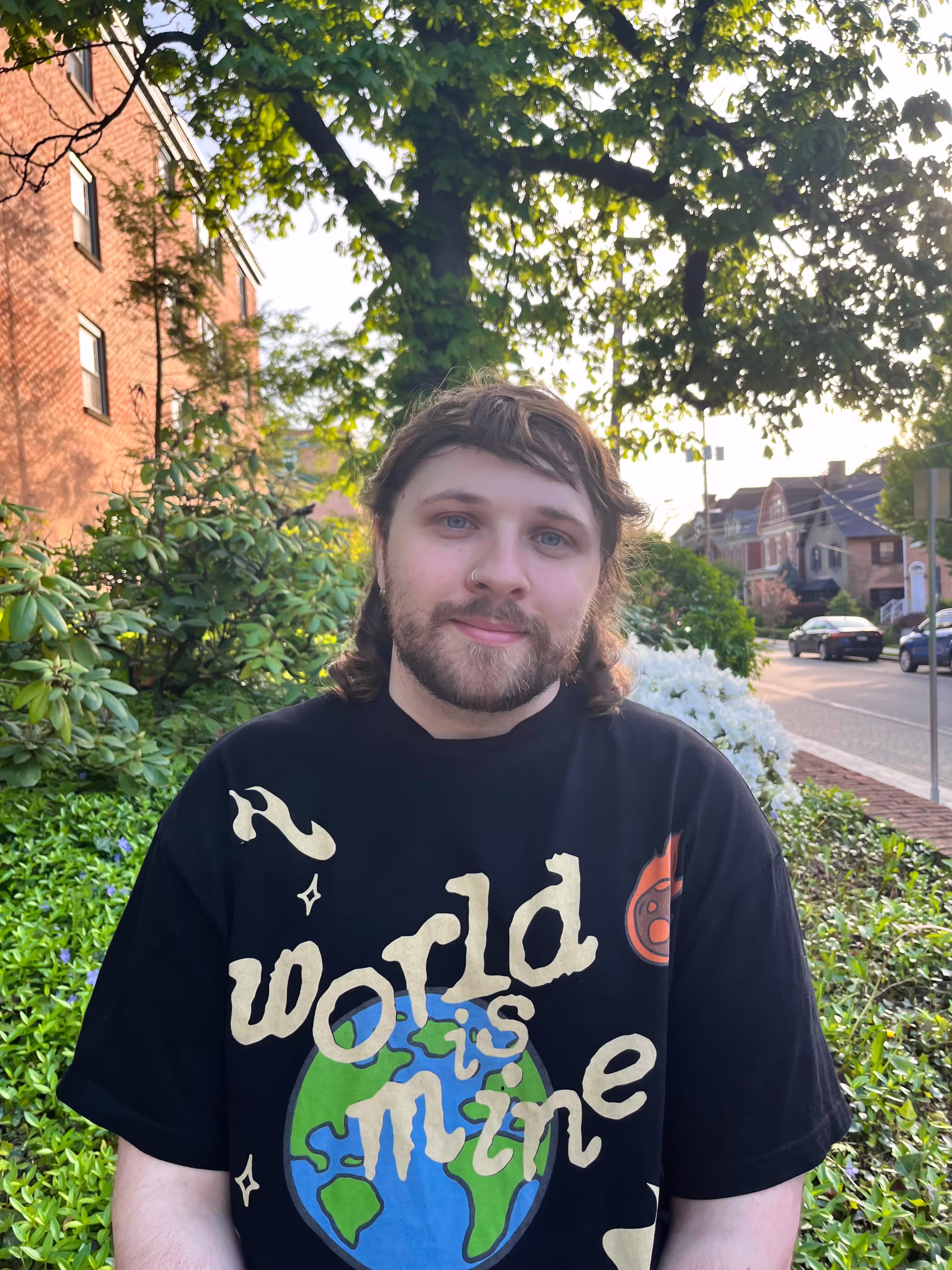 A headshot of Logan LaMaster with a t shirt that says "World Is Mine" there is greenery and trees in the back. 
