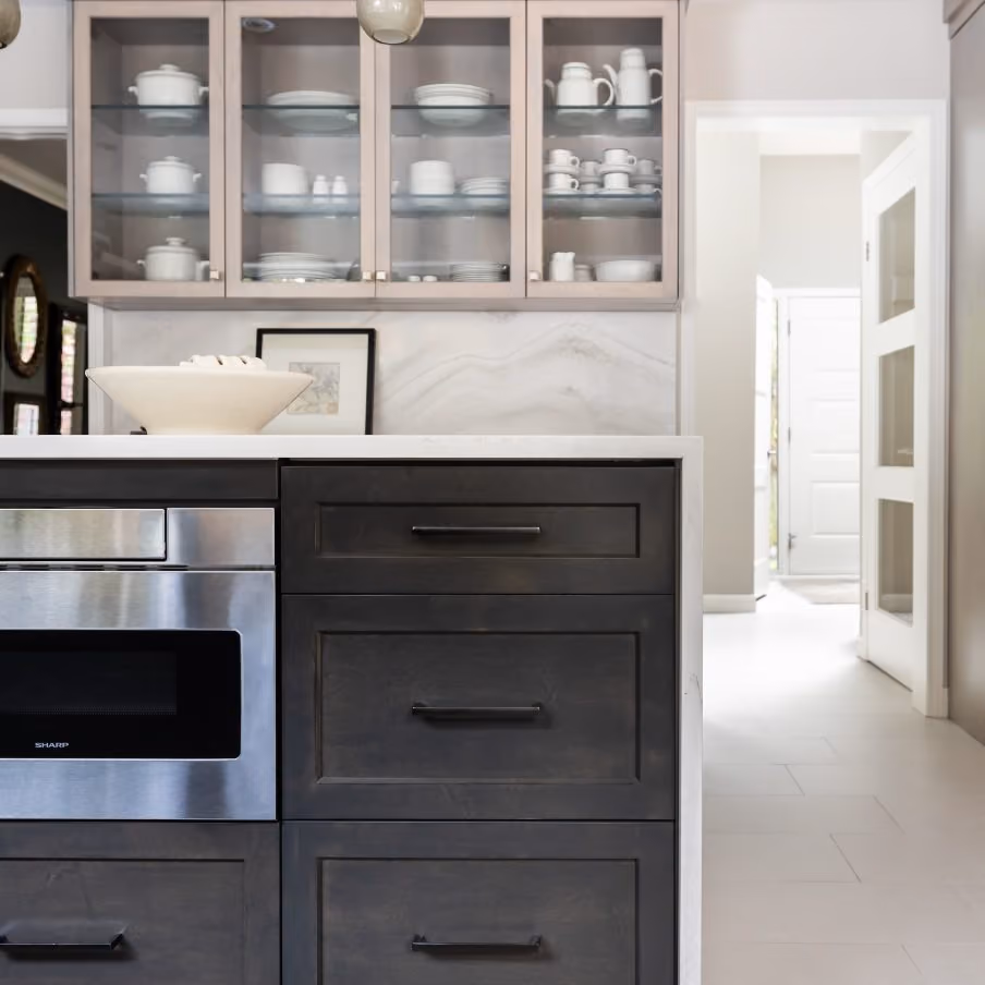 Modern kitchen showing dark wood cabinets, a built-in stainless steel microwave, glass-front upper shelves with white dishes, and a bright hallway in the background.