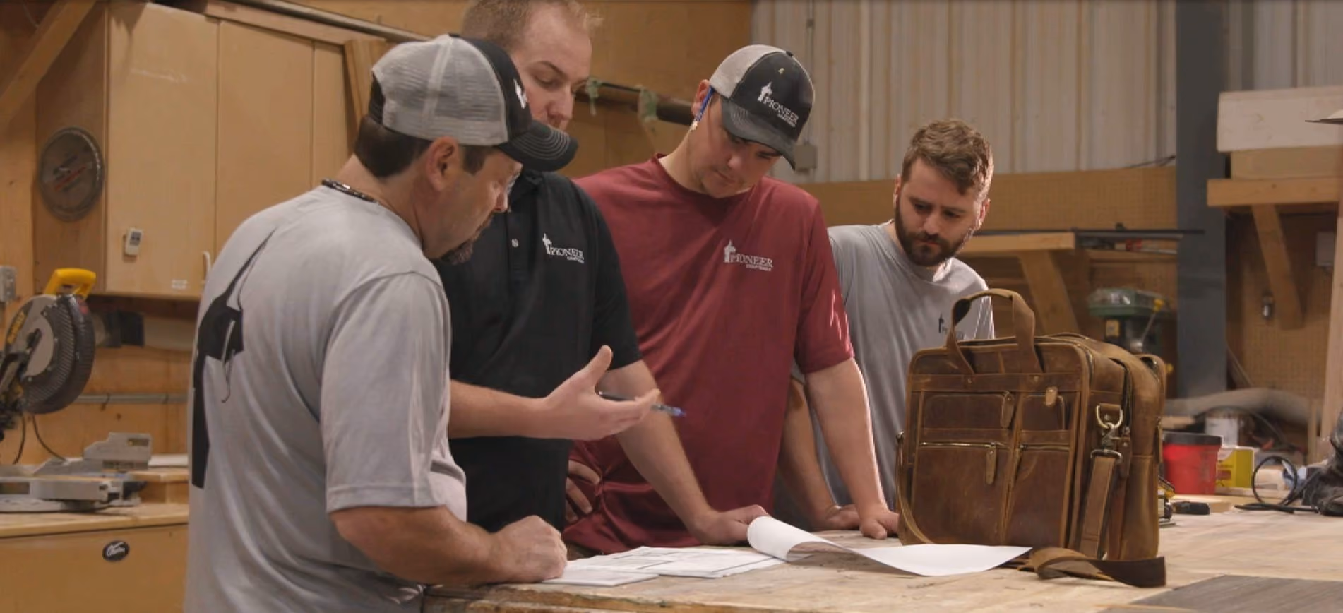 Four men in casual workwear and caps reviewing documents on a wooden table in a workshop with tools and a leather bag nearby.