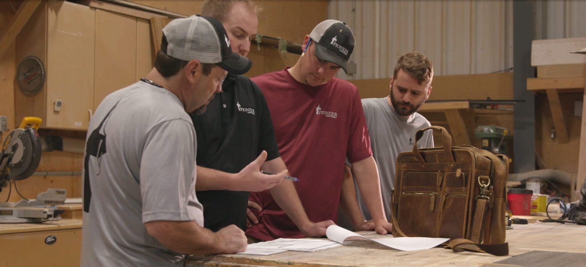 Four men in casual workwear and caps reviewing documents on a wooden table in a workshop with tools and a leather bag nearby.