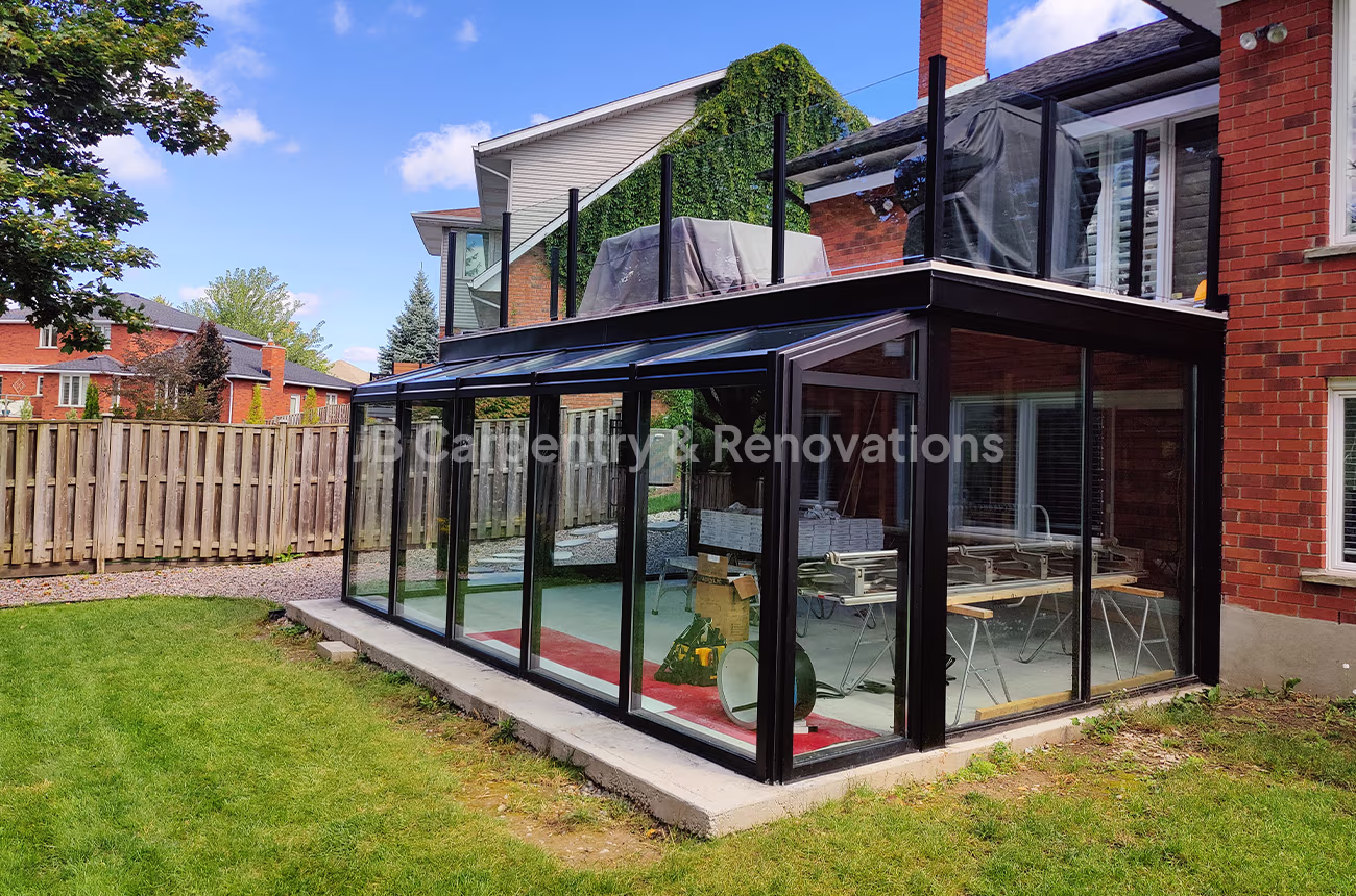 Custom-built glass sunroom addition with upper-level deck and black aluminum framing — exterior renovation by JB Carpentry & Renovations.