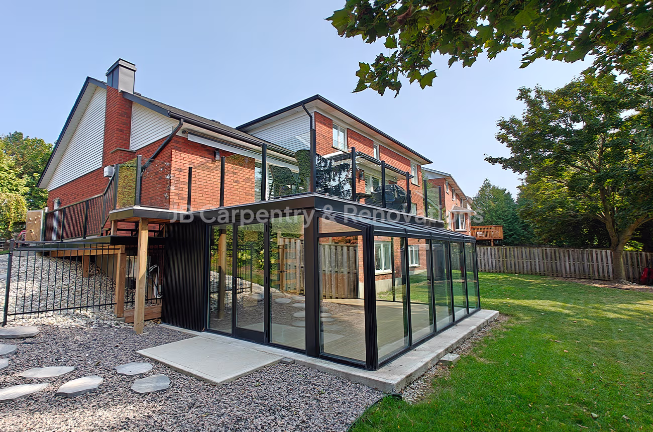 Two-story home renovation featuring a glass sunroom addition with upper-level deck and black aluminum framing — exterior carpentry and design by JB Carpentry & Renovations.