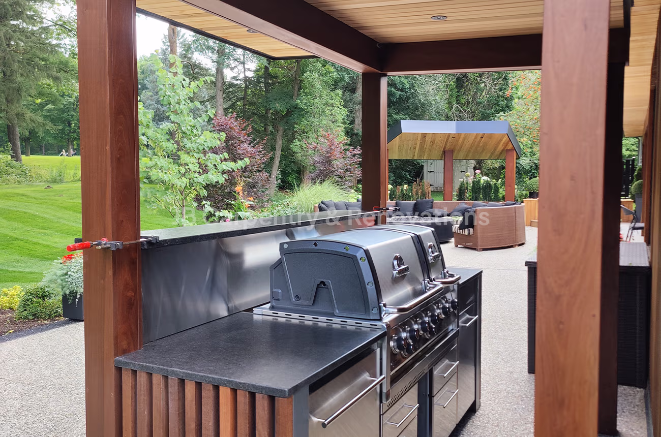 Custom outdoor kitchen with built-in stainless steel grill and wood beam structure, overlooking a modern patio and lounge area — designed and built by JB Carpentry & Renovations.