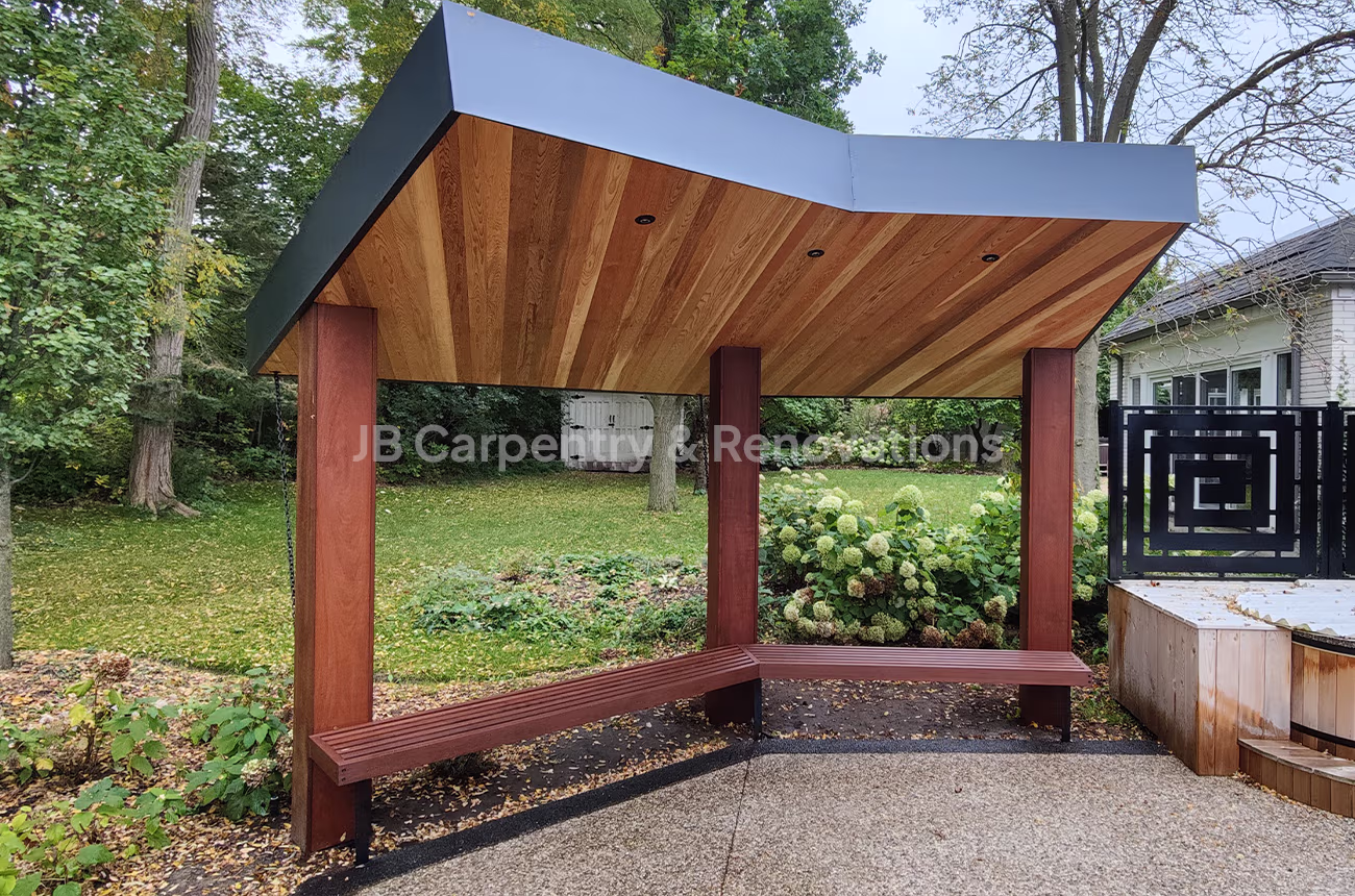 Modern backyard pergola with angled metal roof and wood soffit ceiling, featuring built-in bench seating — designed and constructed by JB Carpentry & Renovations.