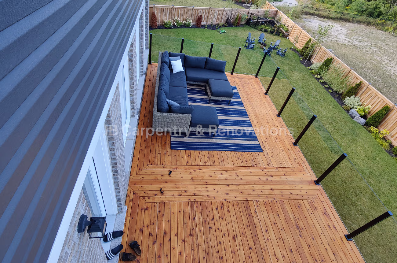 Spacious elevated backyard deck with natural wood finish, black glass railing posts, and outdoor lounge furniture — custom-built by JB Carpentry & Renovations.