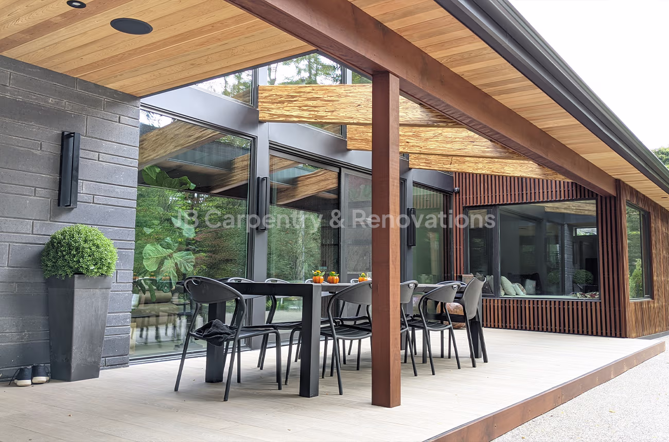 Modern covered patio with exposed beams, wood soffit ceiling, and black-framed glass doors — outdoor dining space by JB Carpentry & Renovations.