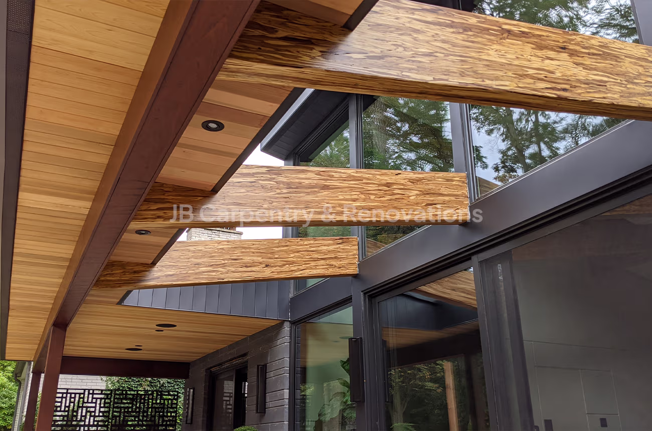 Modern covered patio with exposed beams, wood soffit ceiling, and black-framed glass doors — outdoor dining space by JB Carpentry & Renovations.