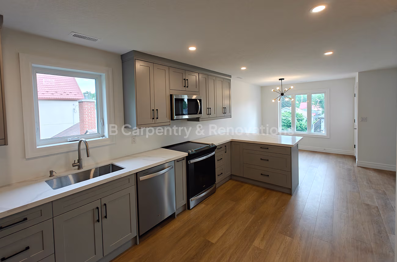 L-shaped kitchen renovation with soft grey shaker cabinets, quartz countertops, and stainless steel appliances by JB Carpentry & Renovations.