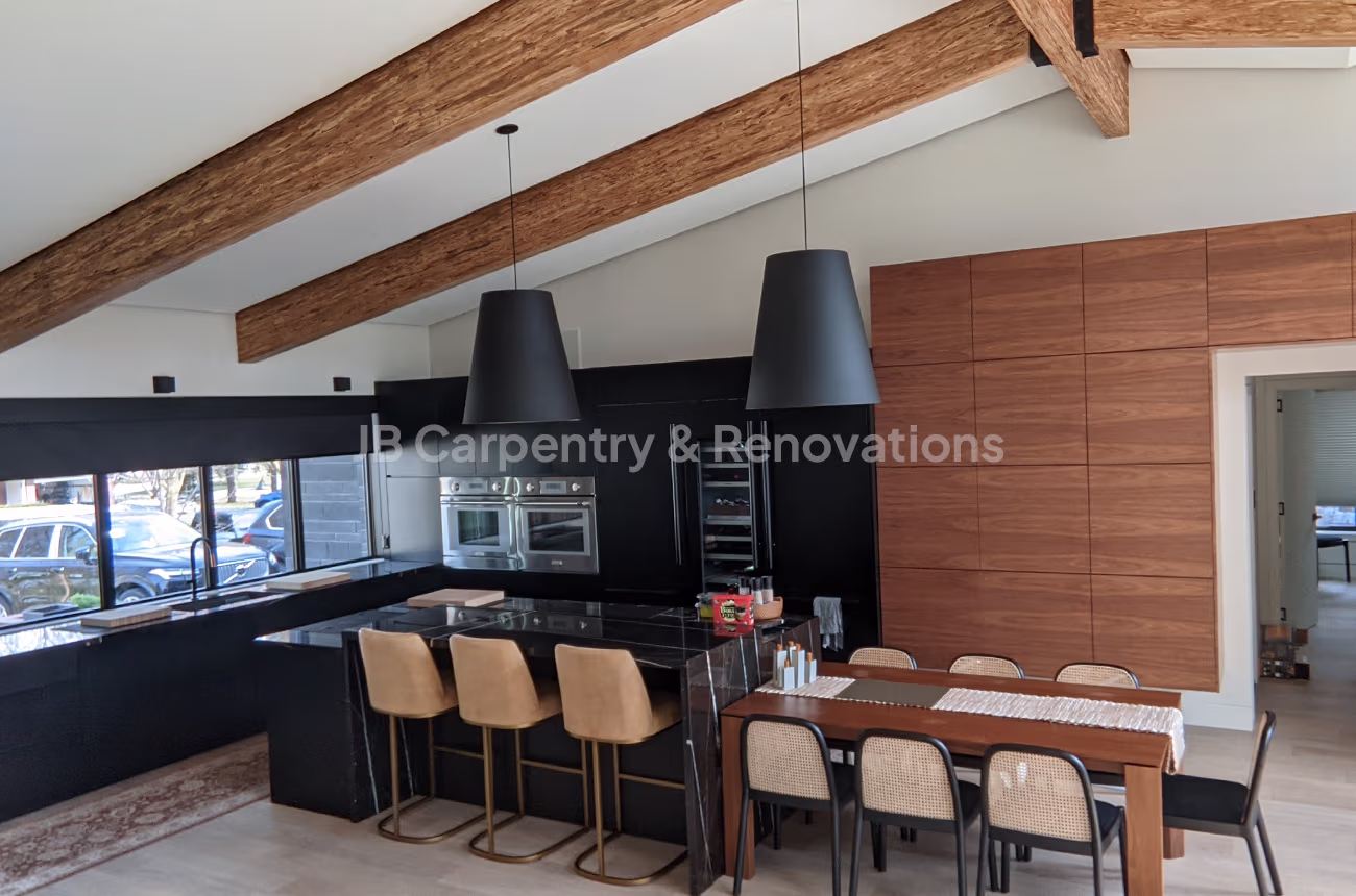 Modern architectural kitchen with exposed beams, wood cabinetry, black countertops, and pendant lighting — JB Carpentry & Renovations.