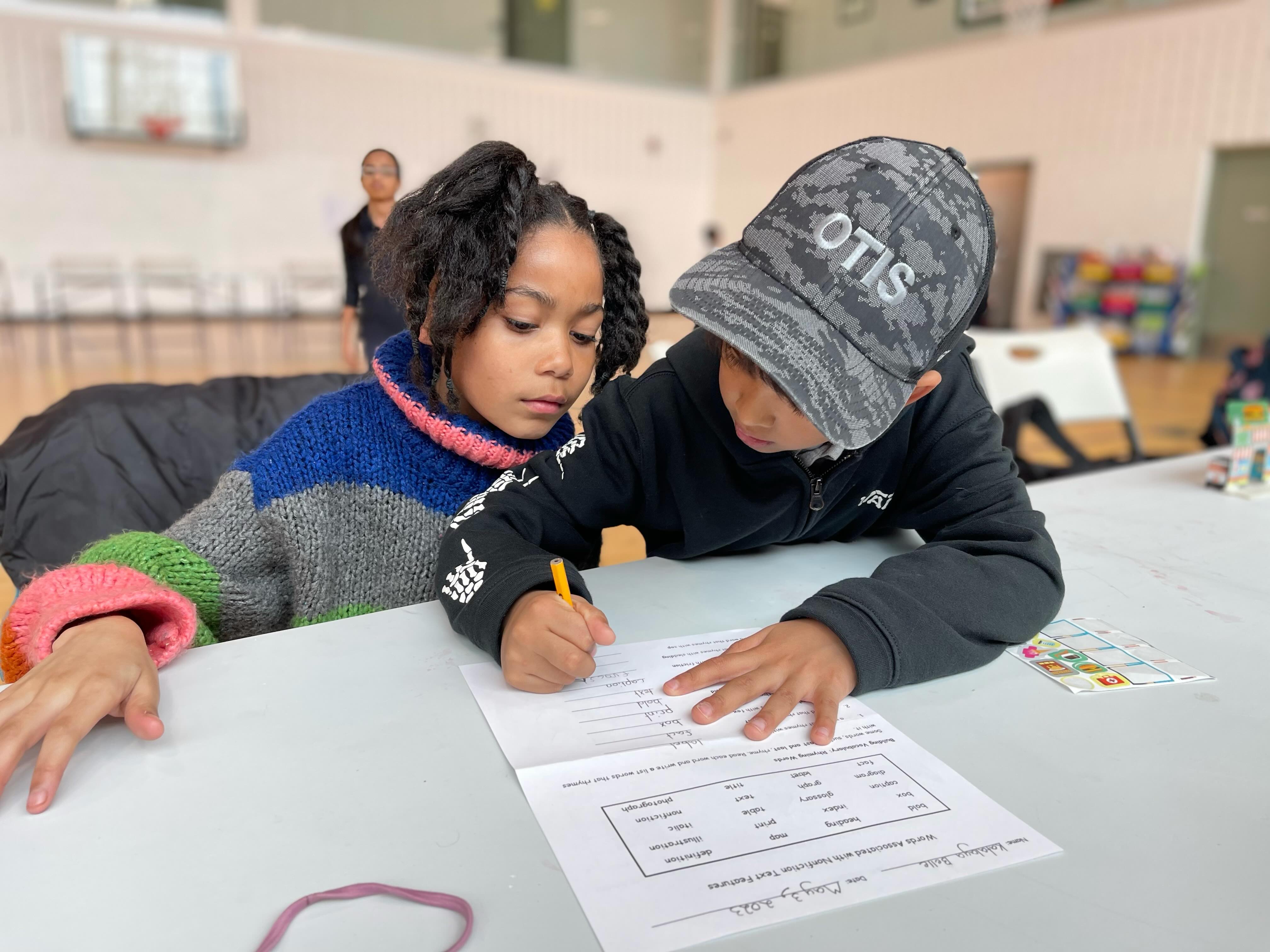 Two children working together on a worksheet at a table in a gymnasium.