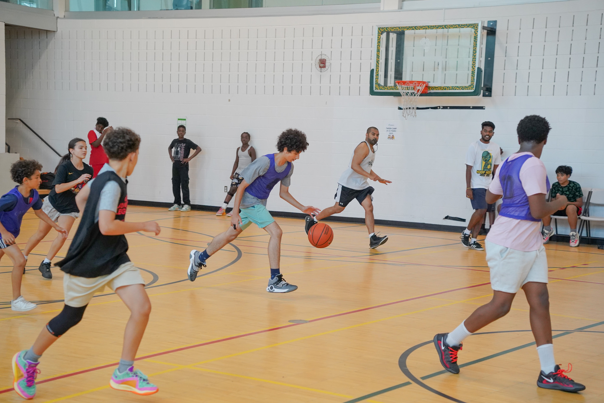 Indoor basketball game with kids and adults. A young player dribbles while others, in purple jerseys, defend. Spectators watch from the sidelines. Energetic scene.