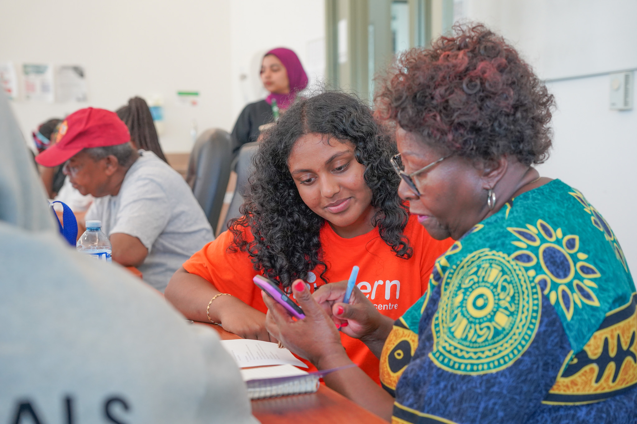 A young woman in an orange shirt helps an older woman with a phone at a community center. They look focused and engaged in a learning atmosphere.