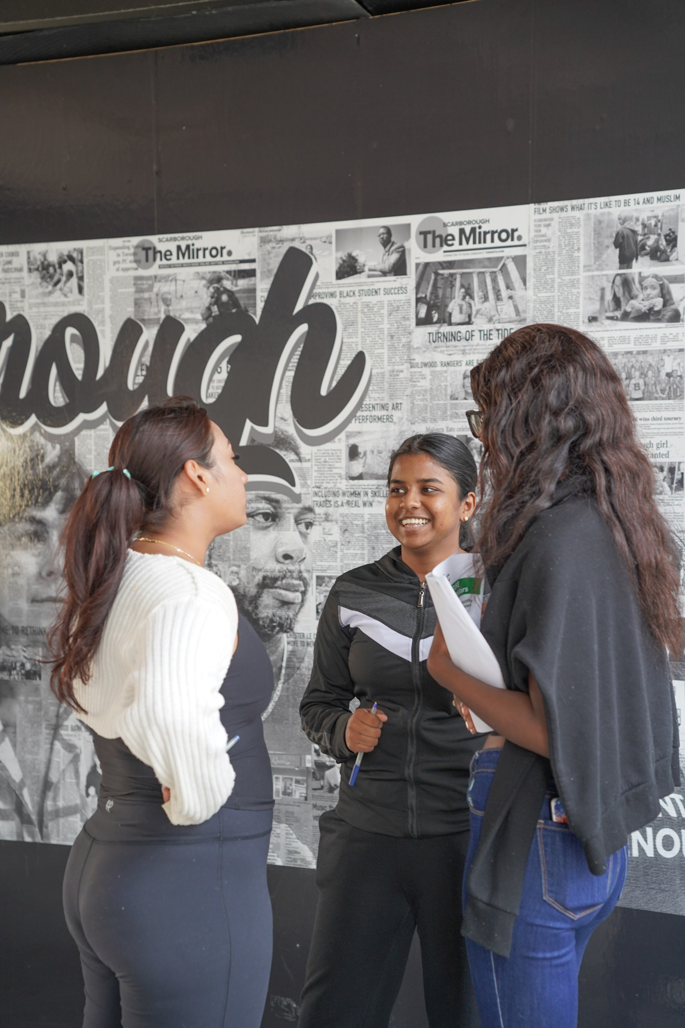 Three women engaged in conversation in front of a wall. They appear focused and animated.