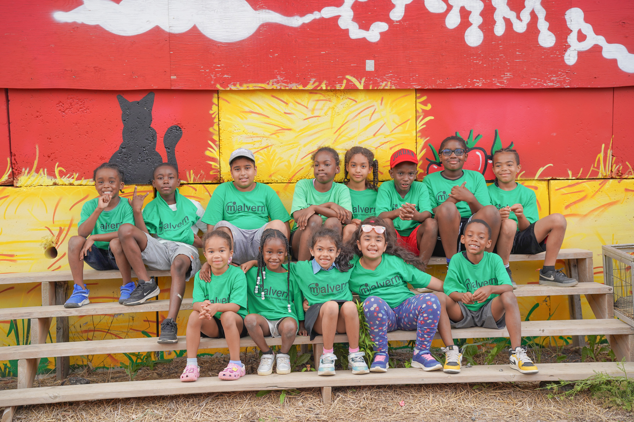 Group of children wearing green Malvern Family Resource Centre shirts sitting on wooden bleachers with a colorful mural background.
