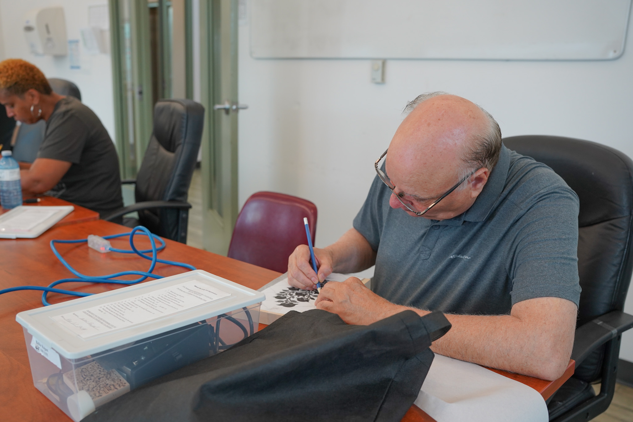 Elderly man concentrating on drawing with a pencil at a table in a classroom setting.