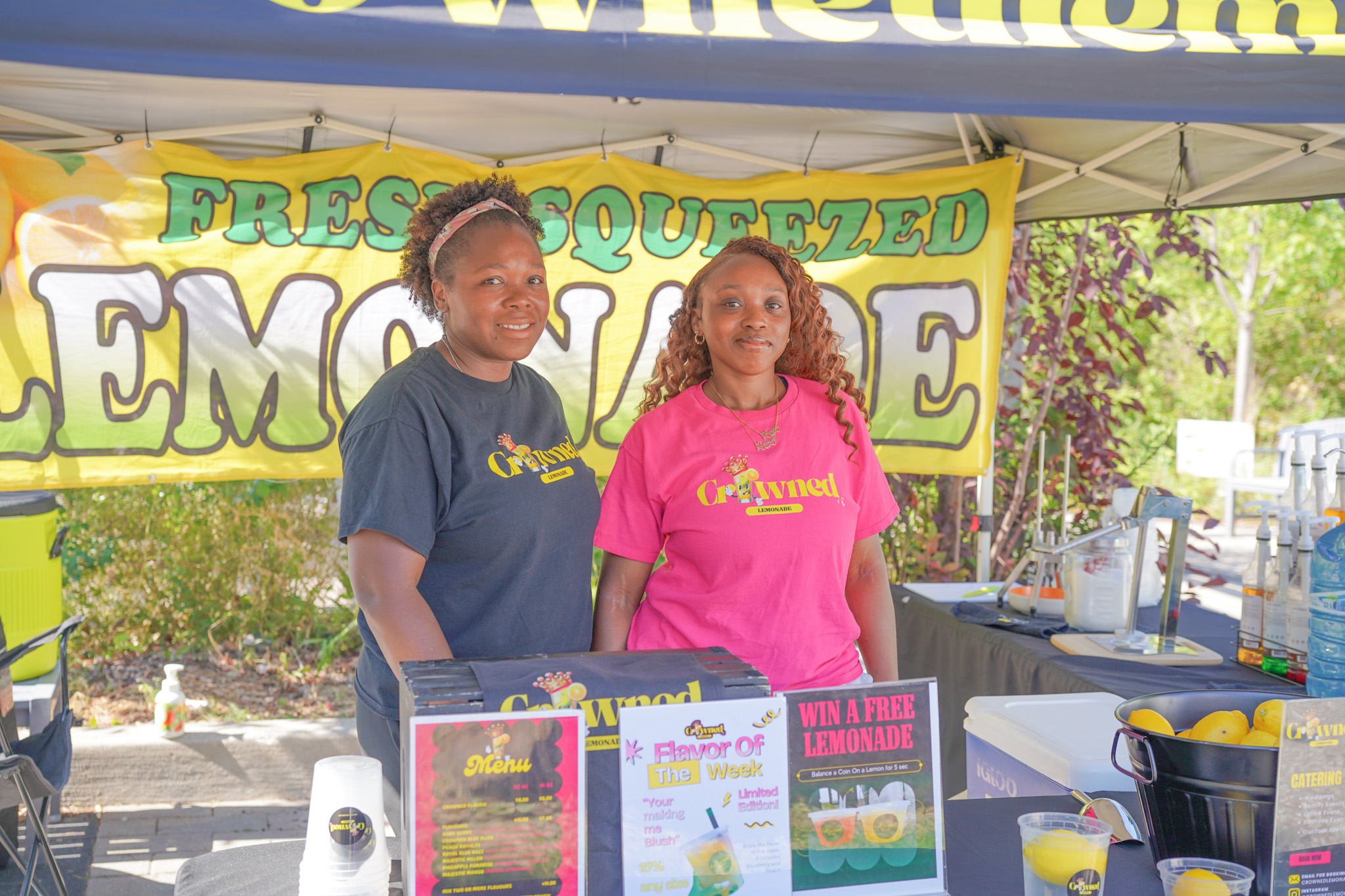 Two women standing behind a lemonade stand with signs advertising fresh squeezed lemonade, flavor of the week, and a chance to win free lemonade.