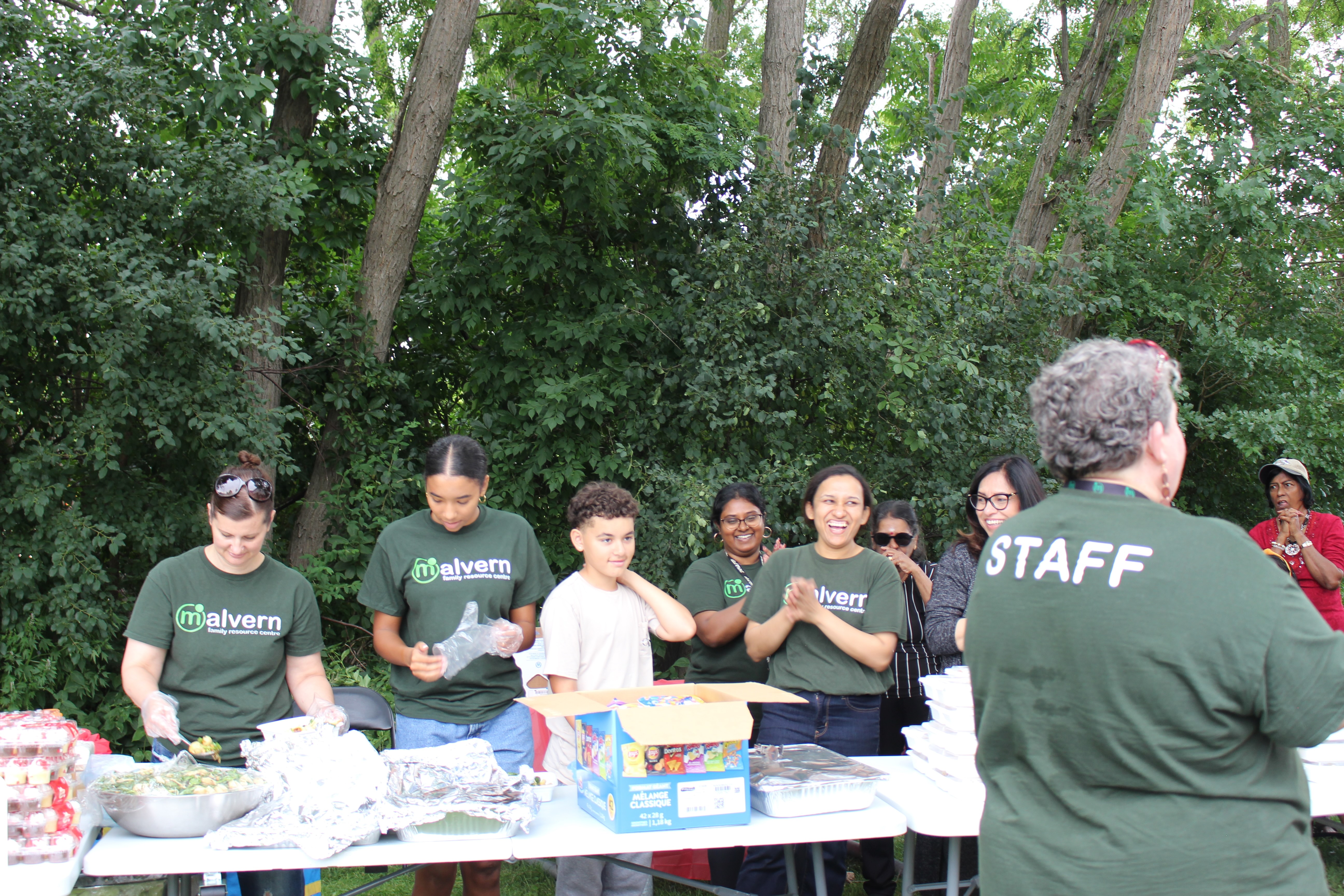 Group of people, some wearing Malvern Family Resource Centre shirts, serving food outdoors at a table with trees in the background.