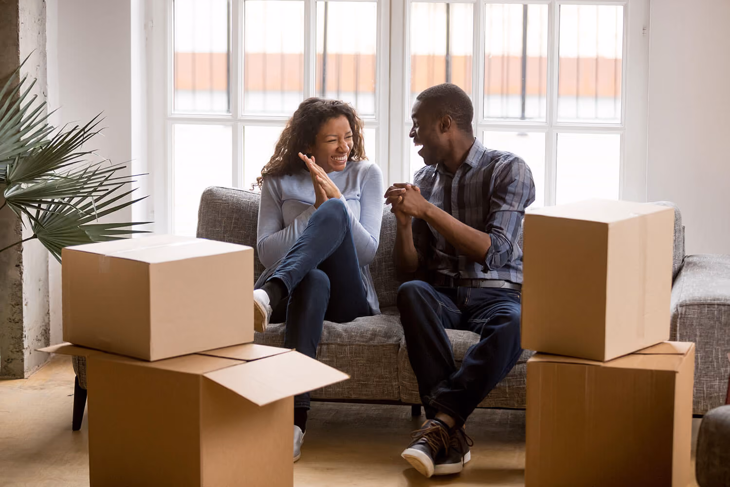 A happy couple sitting on a couch, smiling and talking to each other, surrounded by cardboard moving boxes in a bright room.