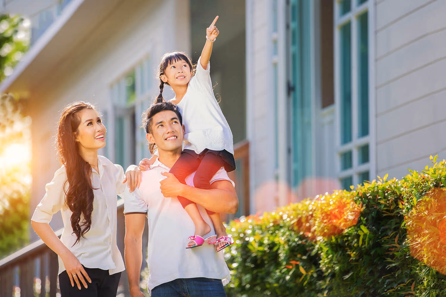 A smiling father carrying his young daughter on his shoulders outdoors. The daughter is looking up and excitedly pointing at something in the distance.