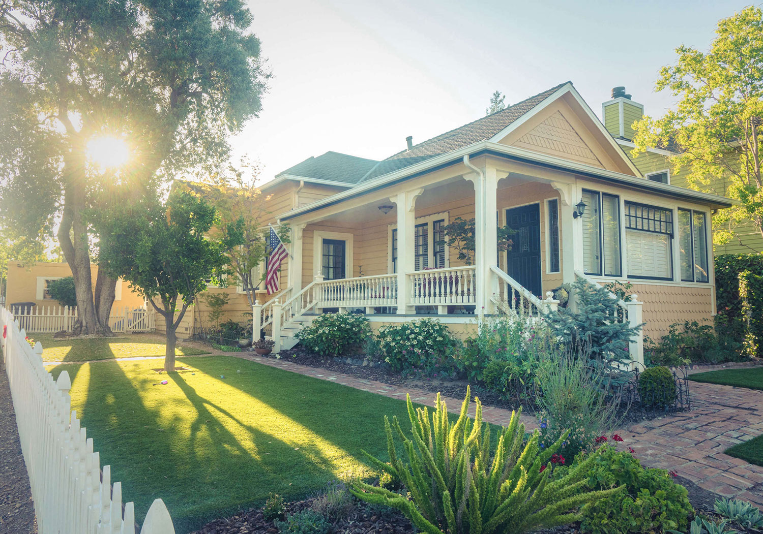 Yellow home with white picket fence & cobblestone brick on a sunny day. The Home Buyers, real estate agents serving Minnesota.